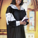 A  senior-adult woman in her black and white pastoral robe inside an old English Gothic church.